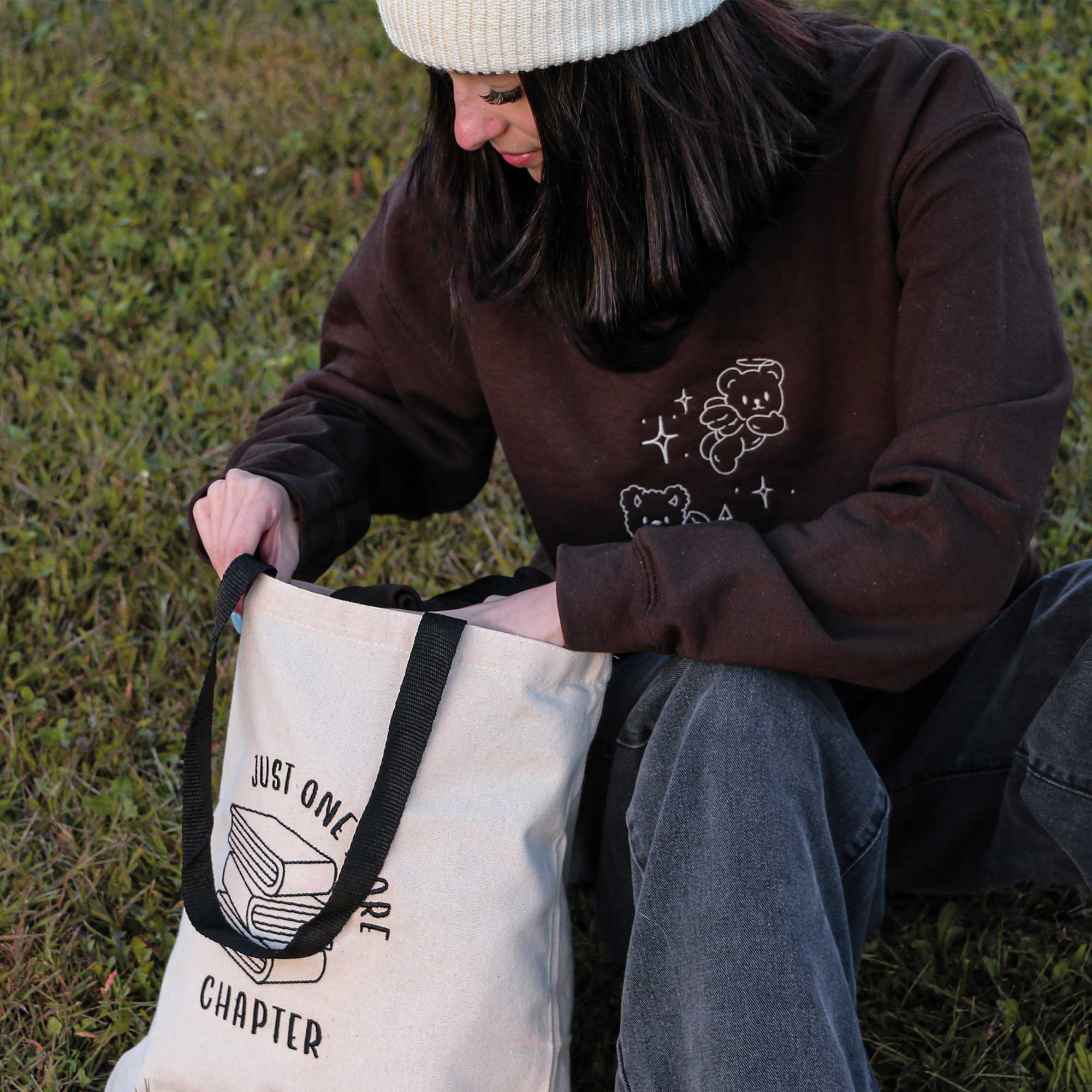 jeune femme portant un crewneck brun teddy, un tote bag one more chapter avec une pile de livres de lecture