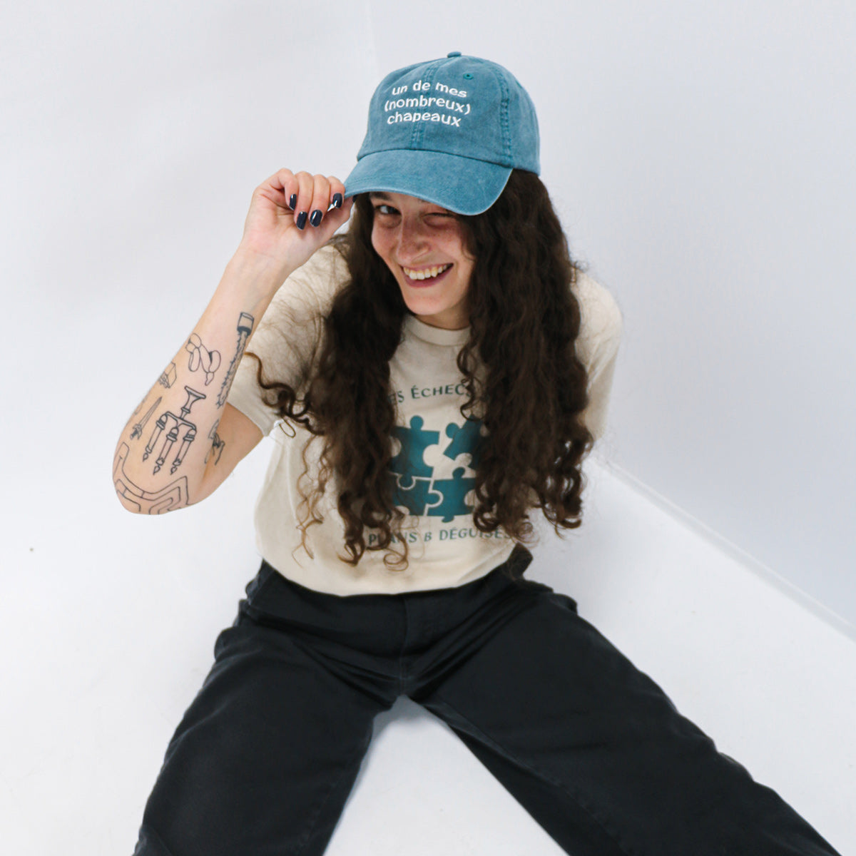 photo lifestyle en studio de photographie d'une jeune femme aux cheveux frisés bruns foncés portant une casquette turquoise délavée avec une broderie disant "un de mes nombreux chapeaux". Design en collaboration avec l'ACEE