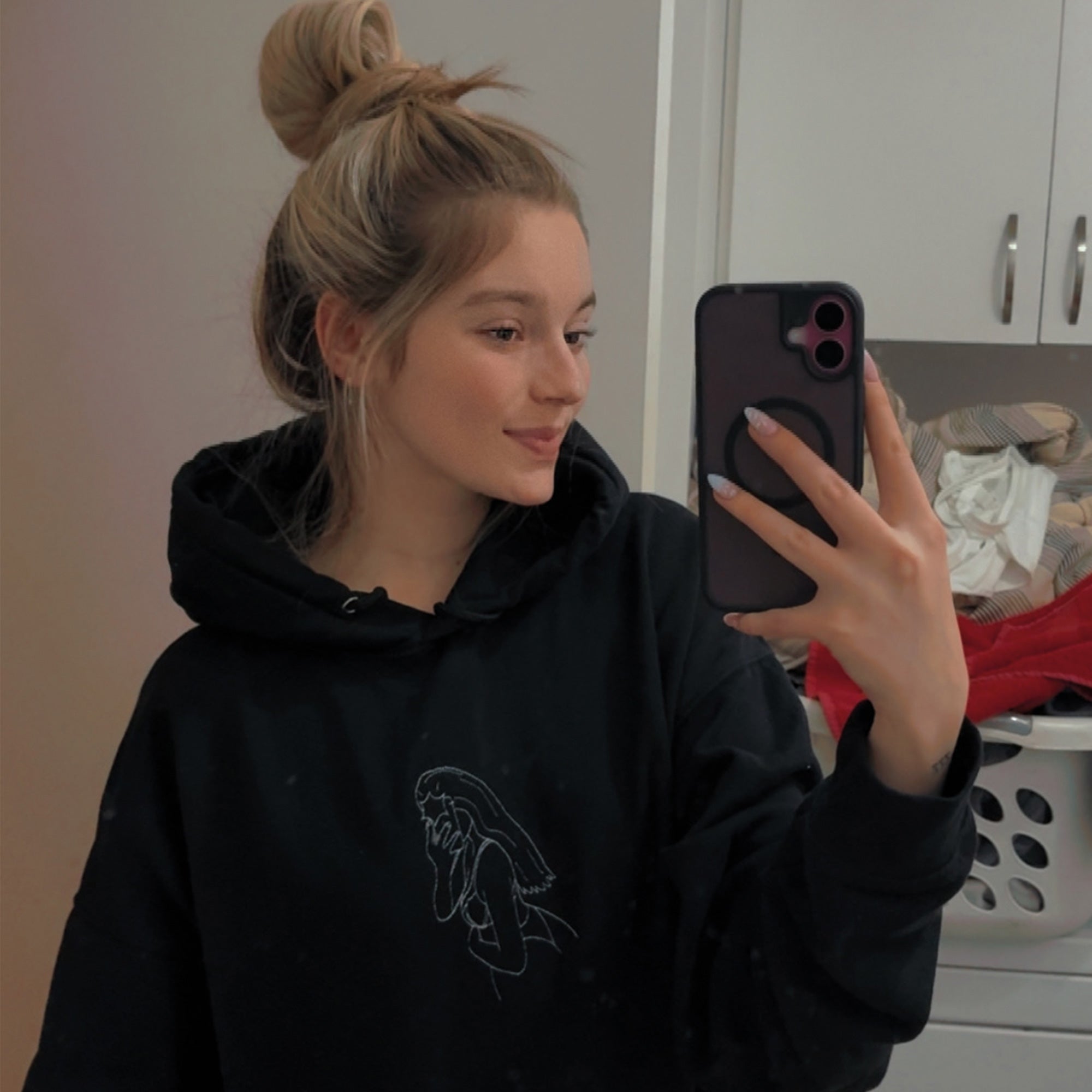 photo lifestyle dans le miroir d'une jeune femme portant un hoodie à capuchon noir avec une silhouette coquine personnalisée brodée en blanc au centre