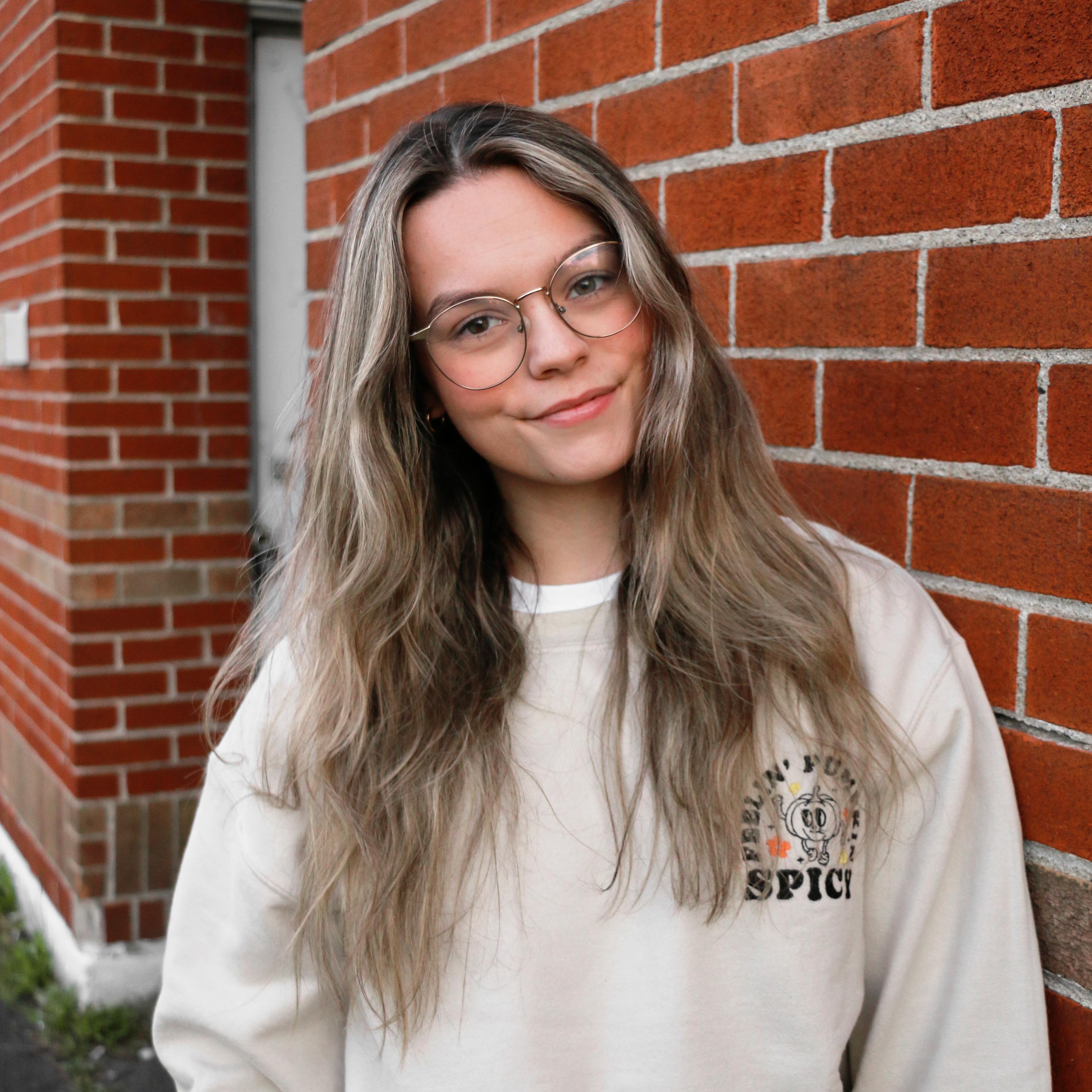 jeune femme aux cheveux longs portant un sweatshirt beige avec une broderie d'automne de citrouille avec l'inscription feeling pumpkin spicy autour avec de petites fleurs jaunes et oranges