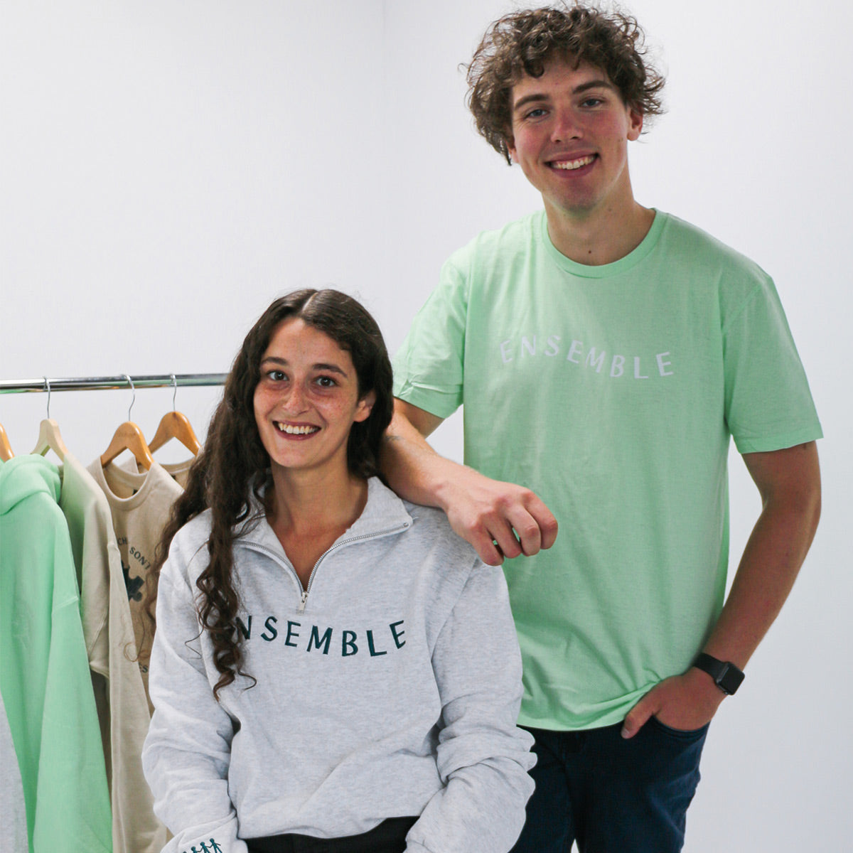 photo lifestyle dans un studio d'une jeune femmes aux cheveux longs bruns frisés portant le quarter zip "ensemble". Un jeune homme est accoté sur elle et il porte le t-shirt menthe ensemble. Les deux items font partie de la collection de Brodame X ACEE.
