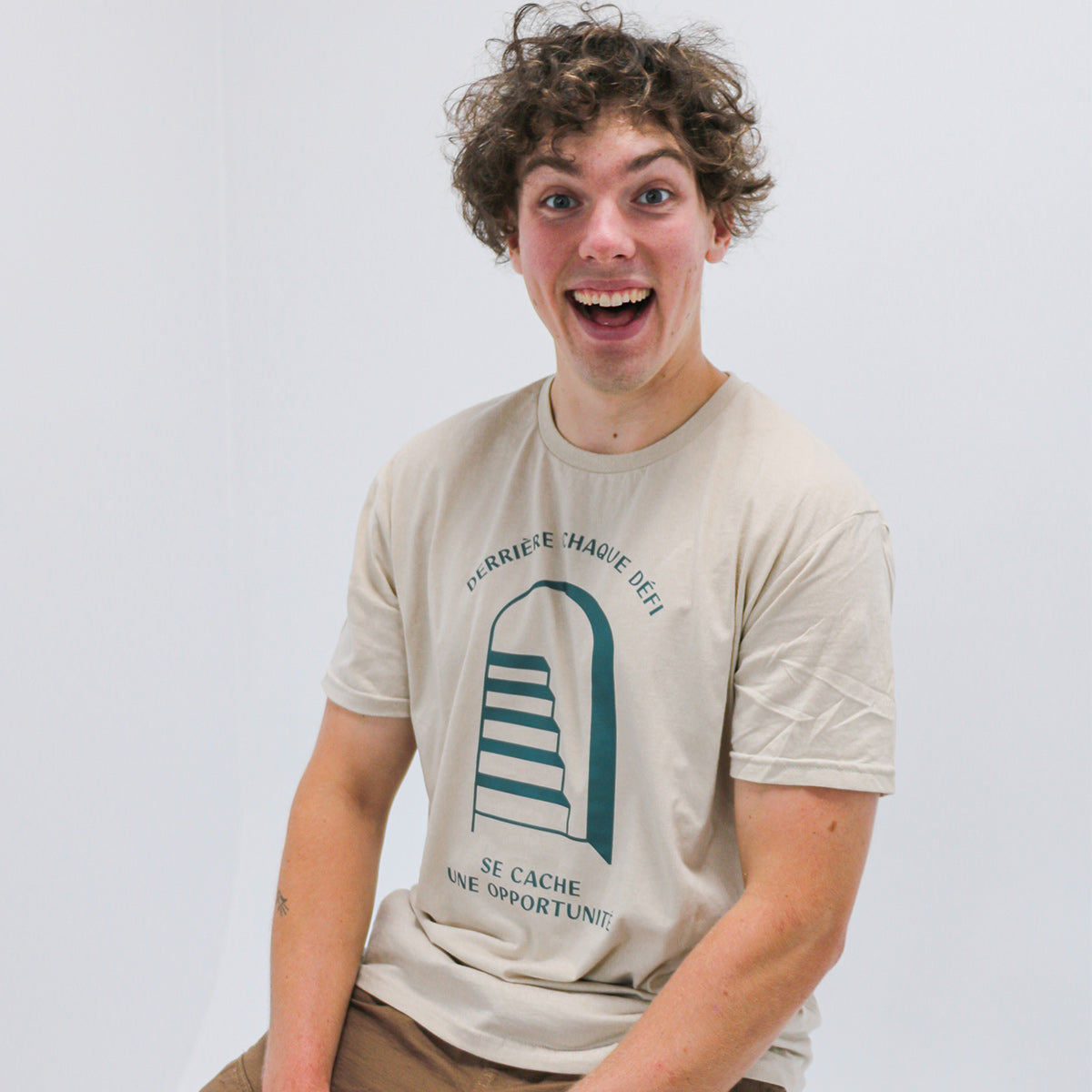 jeune homme aux cheveux frisés bruns souriant portant un t-shirt beige avec un imprimé d'une porte qui s'ouvre révélant des escaliers avec la phrase "derrière chaque défi se cache une oppourtunité"